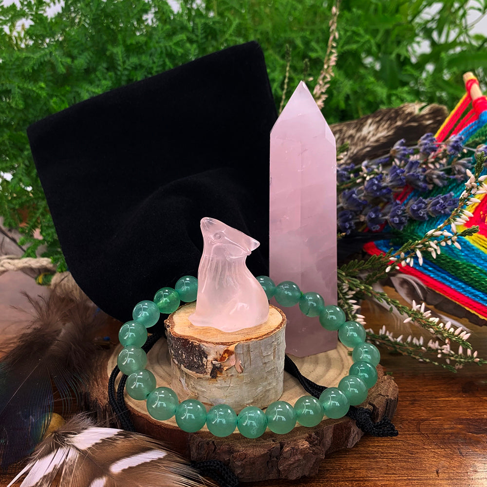 A carved rose quartz wolf on a wood slice, a rose quartz point, a green aventurine bead bracelet and a black velvet pouch together