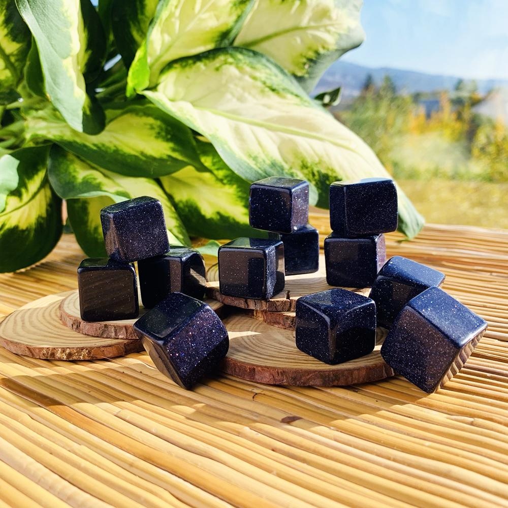 Glossy midnight-blue blue sandstone tumbled cubes flecked with copper sparkle, set on wood slices in sunlight beside a leafy plant