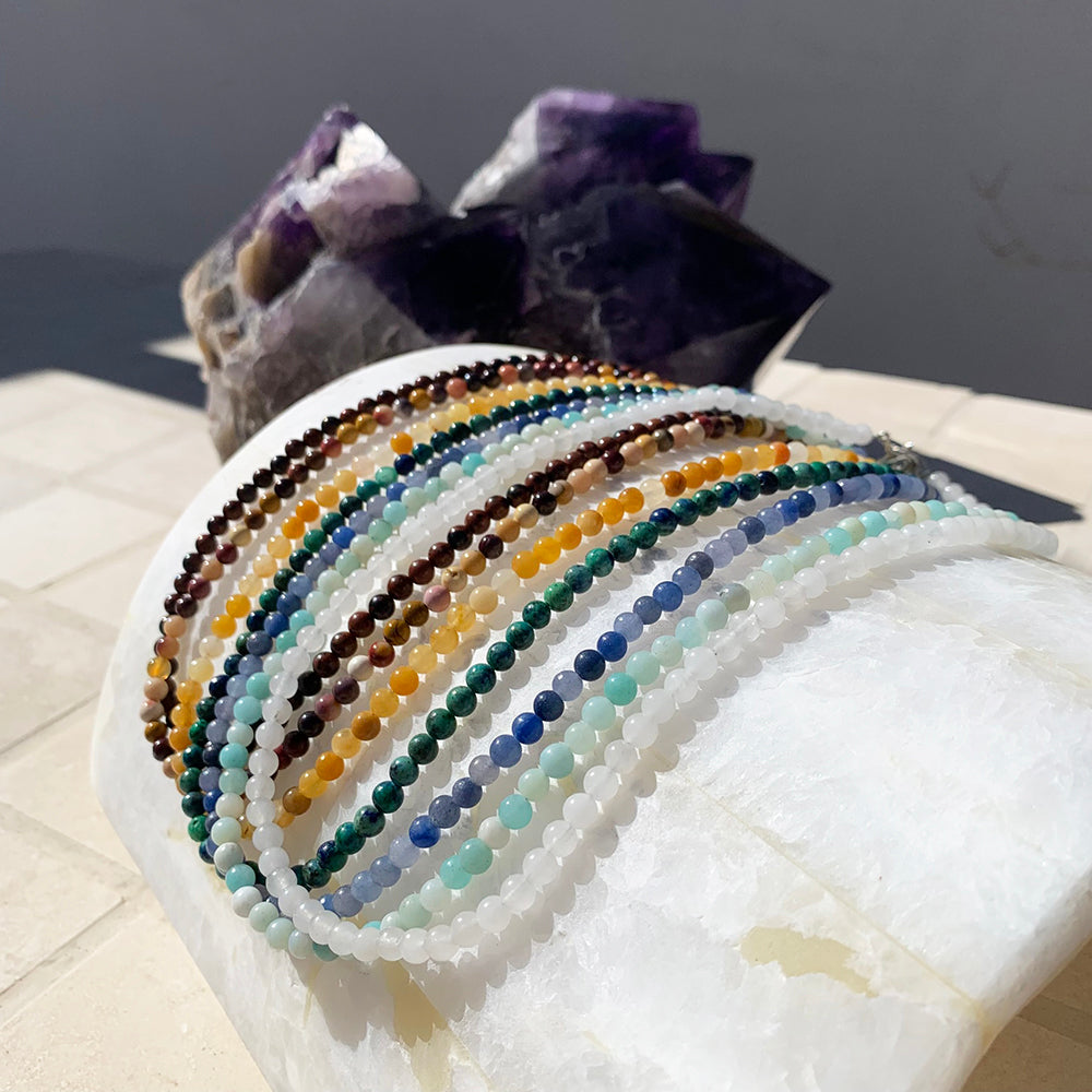 Seven beaded crystal chakra necklaces in red, orange, yellow, green, blue, teal and white draped over a selenite slab in front of amethyst