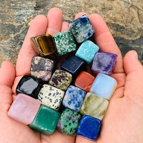Two cupped hands holding nineteen colourful tumbled crystal cubes, including a green aventurine cube