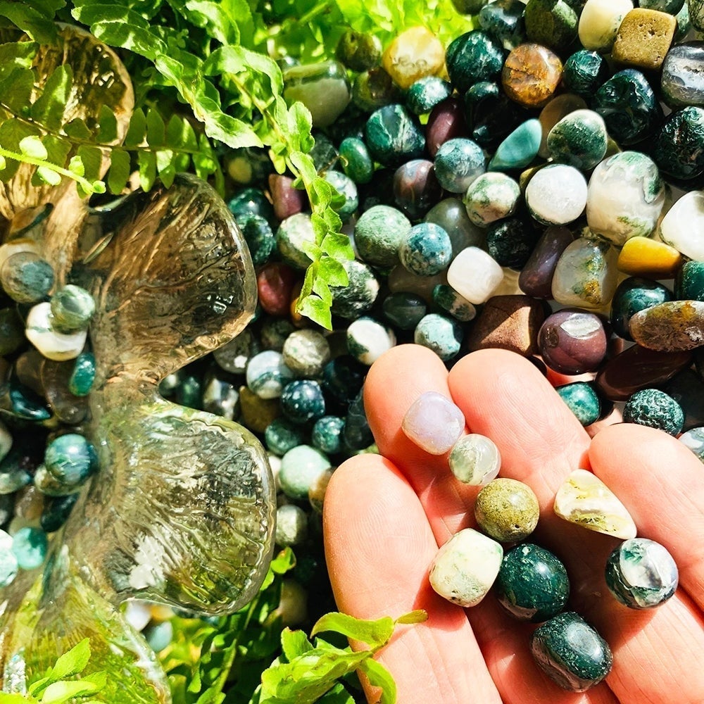 Tumbled green moss agate mini gemstones resting across the fingers of one hand above a heap of more stones and green ferns