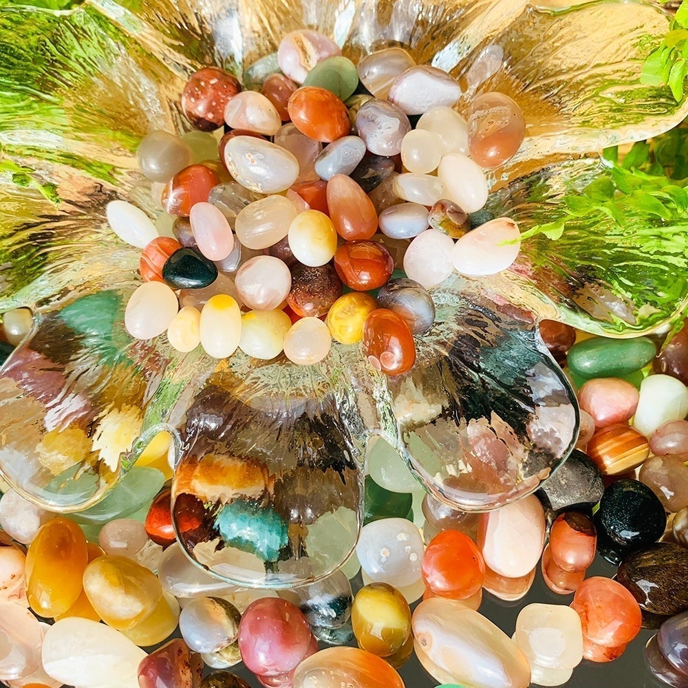 A sunlit heap of polished agate mini gemstones in white, peach, orange, red, green and grey, piled in and around a clear glass dish