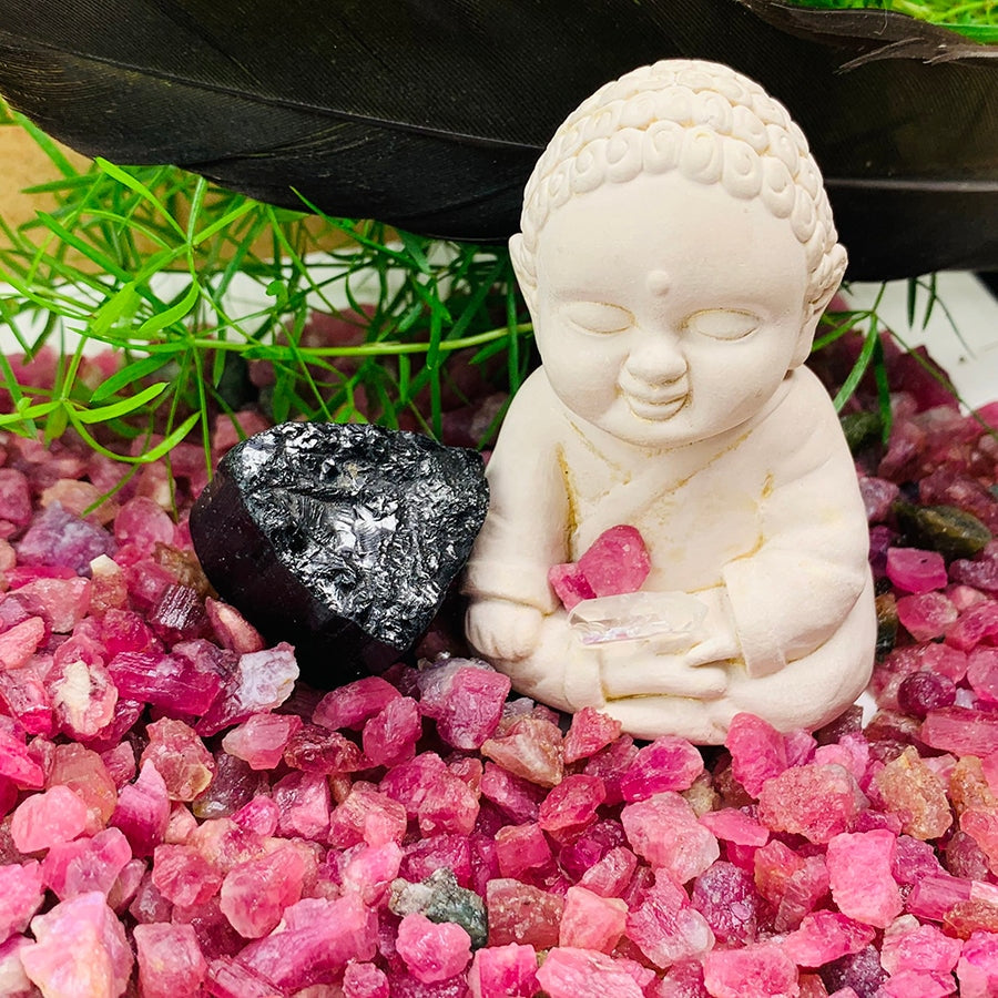 Rough pink tourmaline spread around a white seated figurine and a larger black tourmaline stone, with a black feather and grass behind