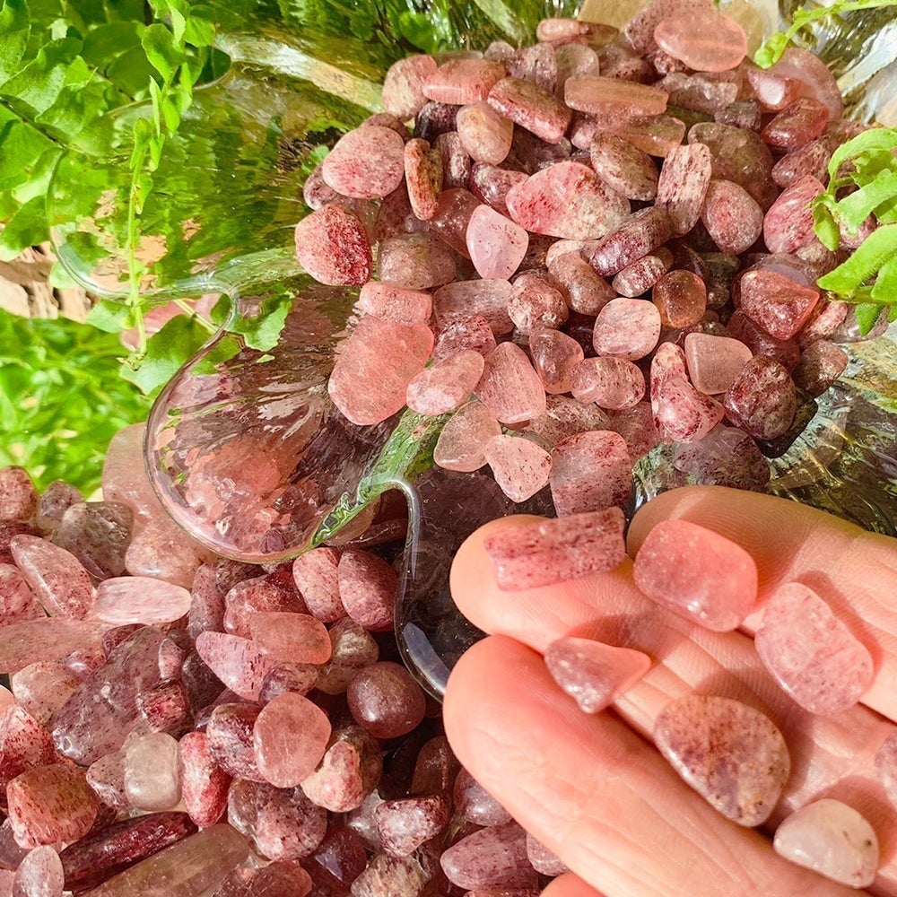 Fingers holding several small tumbled strawberry quartz stones beside a glass bowl overflowing with more